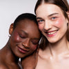 Zwei Frauen mit unterschiedlichen Teints, lehnen sich eng aneinander und tragen den Artistick Blush Stroke von LH Cosmetics.