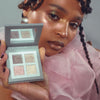 Eine Person in rosafarbenem, durchsichtigem Stoff hält die Aurora-Palette von LH Cosmetics mit vier schimmernden Lidschatten in der Nähe ihres glitzernden Gesichts.