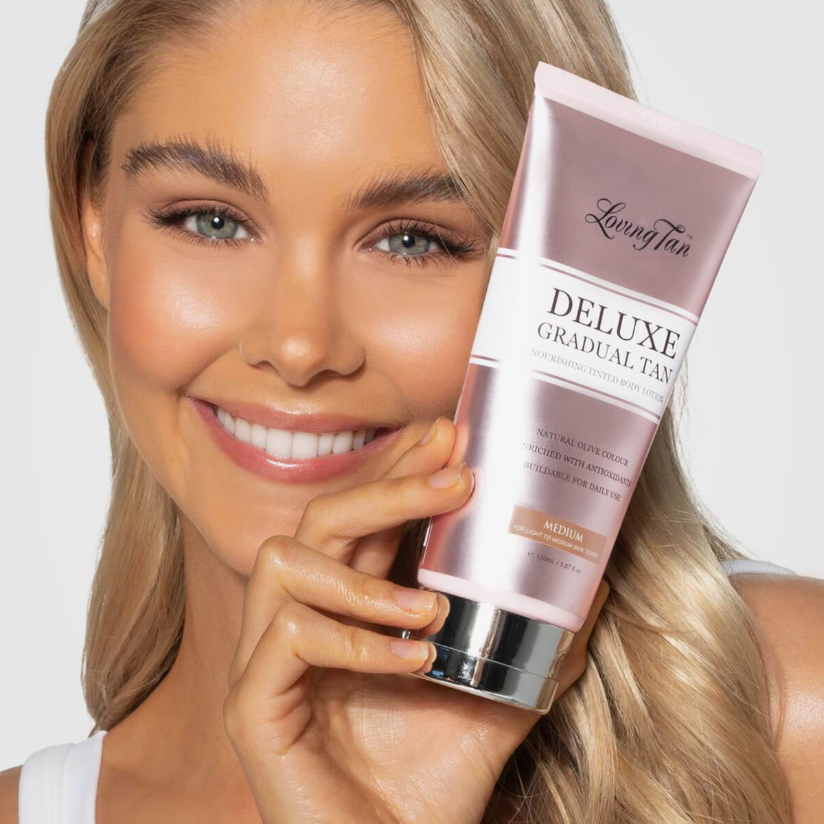 Lächelnde Frau, die eine Tube Deluxe Gradual Tan nah an ihr Gesicht hält.