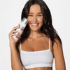 Eine Frau in einem weißen bauchfreien Top lächelt, während sie vor einem schlichten Hintergrund eine Flasche Bronzing Mousse hält.