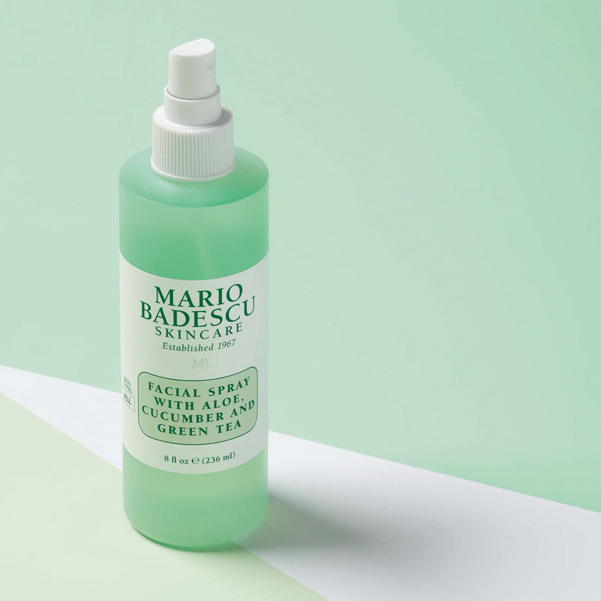 Eine Flasche Mario Badescu Gesichtsspray mit Aloe, Gurke und grünem Tee ist auf einem pastellfarbenen Hintergrund abgebildet.