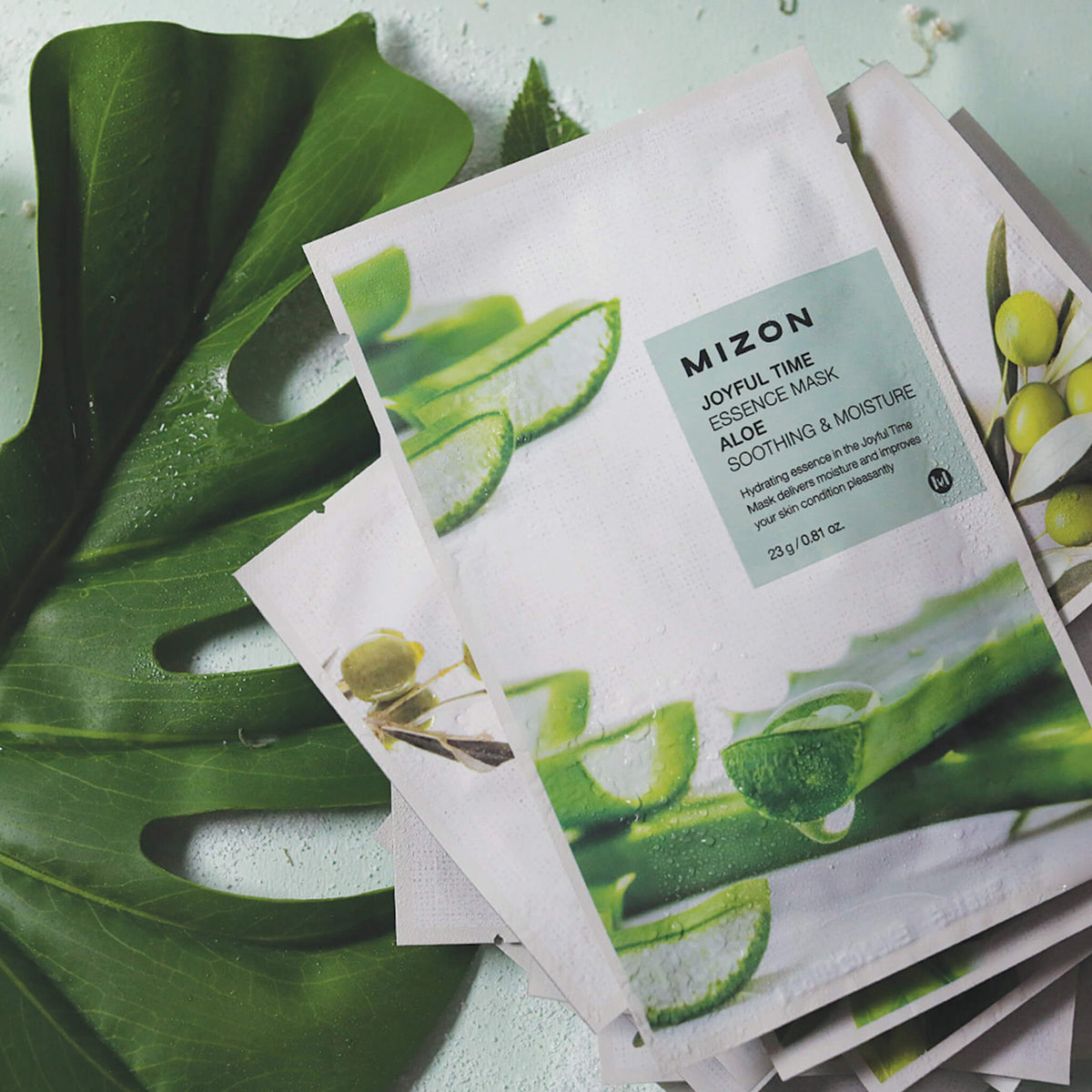 Die MIZON Joyful Time Essence Mask mit Aloe-Design ist auf einem großen grünen Blatt abgebildet, akzentuiert durch Wassertropfen.