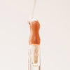 Ein Tropfen Lip Candy Oil in der Farbe Honey gleitet vor einem pfirsichfarbenem Hintergrund über den Applikator.