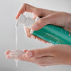 Eine Person gibt Clarion 2 % BHA Cleanser, ein klares Gel aus einer grünen Pumpflasche, vor einem neutralen Hintergrund auf die Hand.