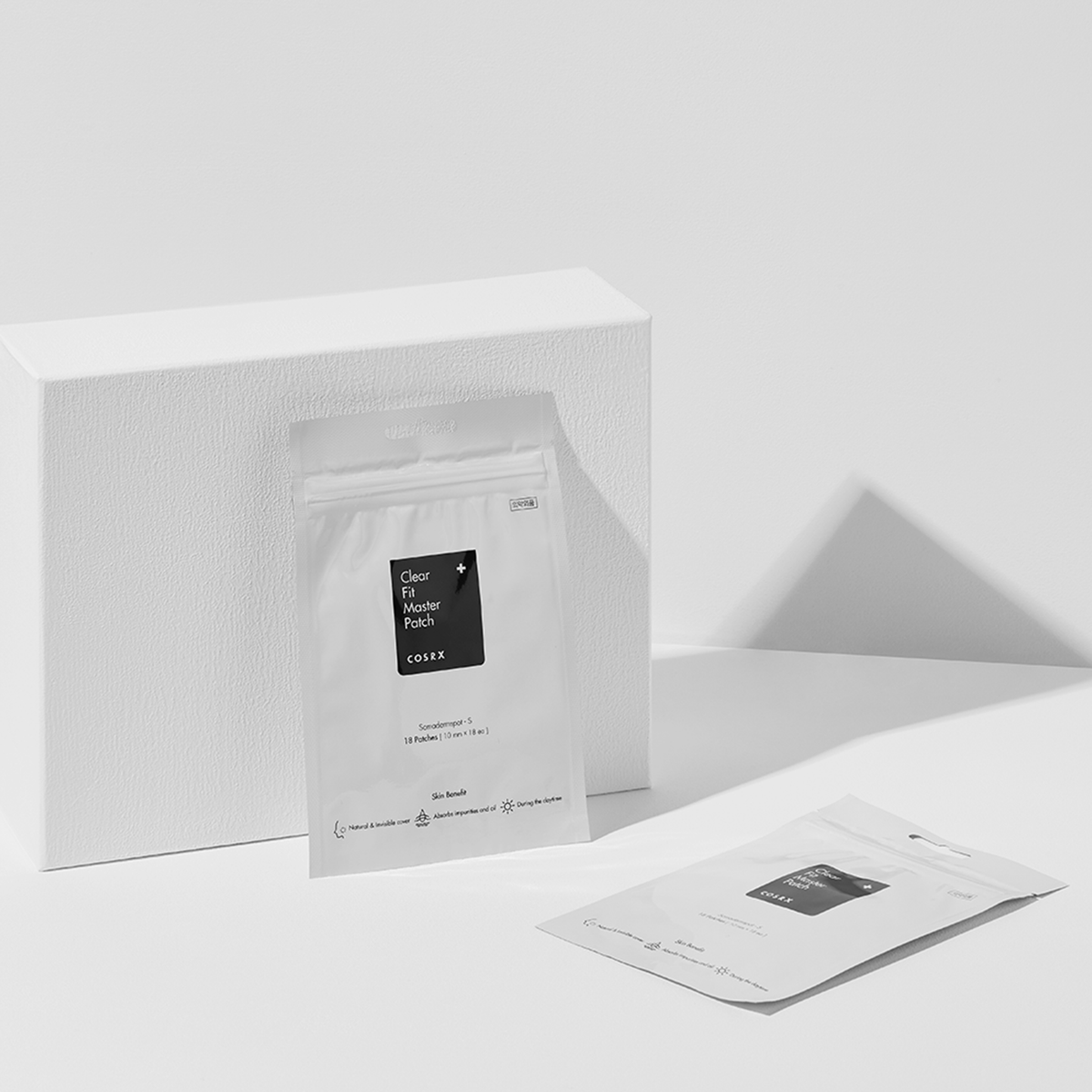 Zwei weiße Clear Fit Master Patch-Verpackugen auf einer weißen Oberfläche mit einem weißen Block im Hintergrund.