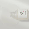 Eine liegende Flasche Glow Deep Serum: Rice + Alpha Arbutin mit heraustretender transparenter Flüssigkeit auf hellem Hintergrund