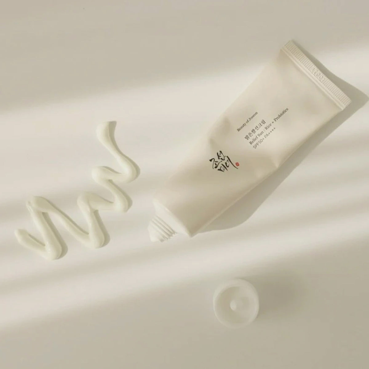 Offene Tube Relief Sun: Rice + Probiotics liegt auf einer hellen Oberfläche, daneben wirft ein cremefarbener Swatch der Lotion