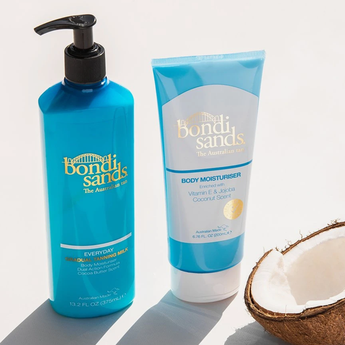 Die Everyday Gradual Tanning Milk (375 ml) und der Body Moisturizer von Bondi Sands stehen auf einer weißen Fläche neben einer halbierten Kokosnuss