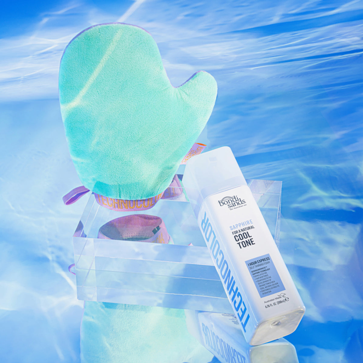 Ein mintgrüner Bräunungshandschuh und eine Flasche Technocolor 1 Hour Express Self Tanning Foam Sapphire auf einem wasserblauen Hintergrund.