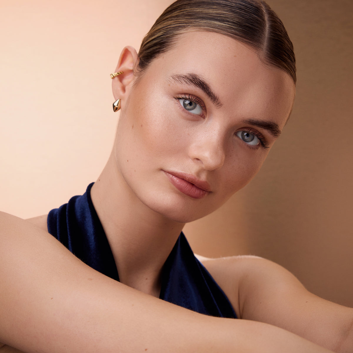 Eine Person mit zurückgekämmtem Haar, einem dunklen Oberteil und goldenen Ohrringen posiert vor einem sanften beigen Hintergrund, hervorgehoben durch den Wonder Woman Butter Bronzer Stick.