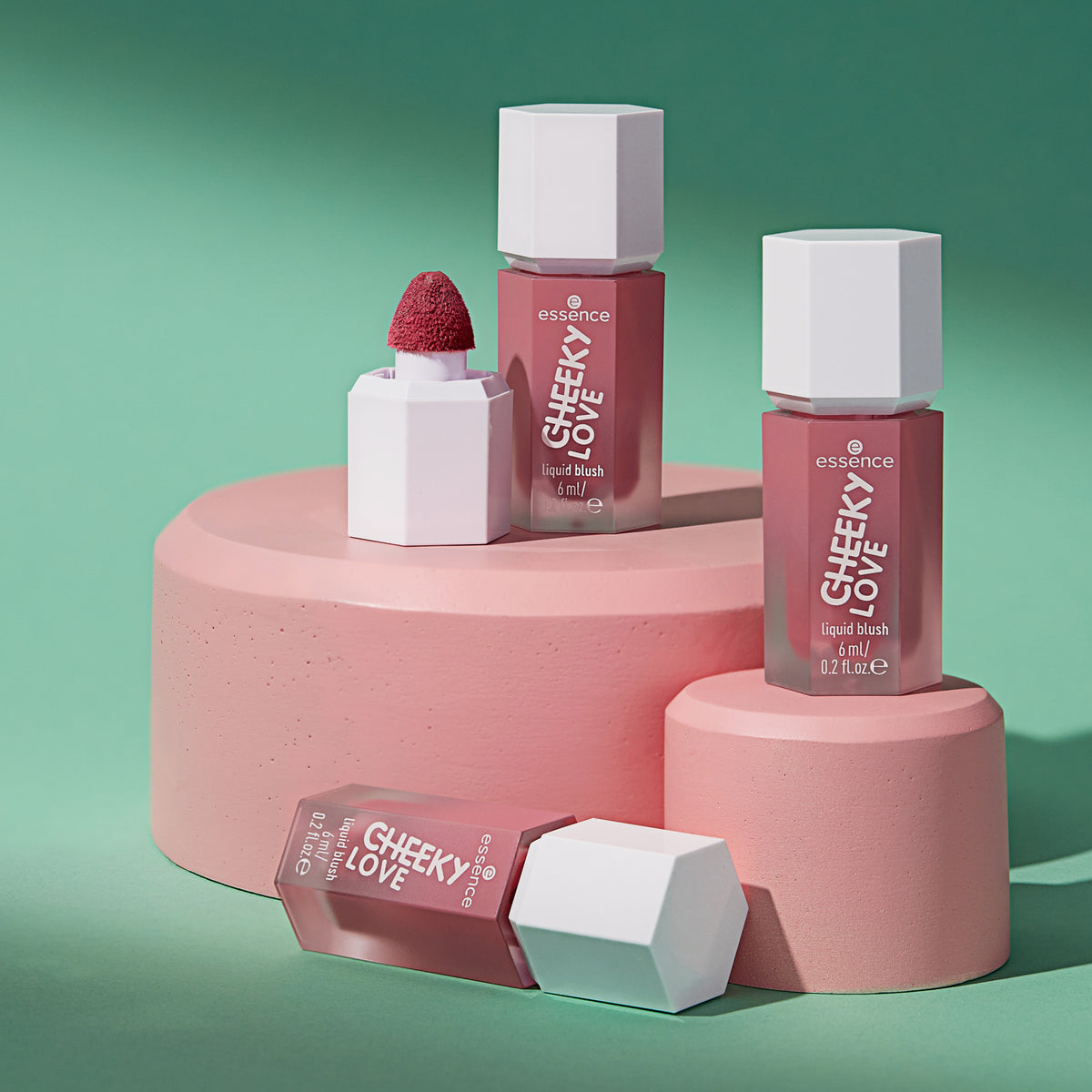 Drei Flaschen CHEEKY LOVE Liquid Blush, präsentiert auf rosa Podesten mit grünem Hintergrund.