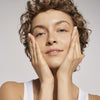 Eine lächelnde Person mit kurzen lockigen Haaren trägt den Vegan Collagen + Organic Oat Barrier & Hydro Toner mit beiden Händen auf ihr Gesicht.