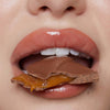 Nahaufnahme von Lippen mit Plumpy Lip Gloss Toffee Brown, die in einen mit Karamell gefülltes Schokostück beißen.