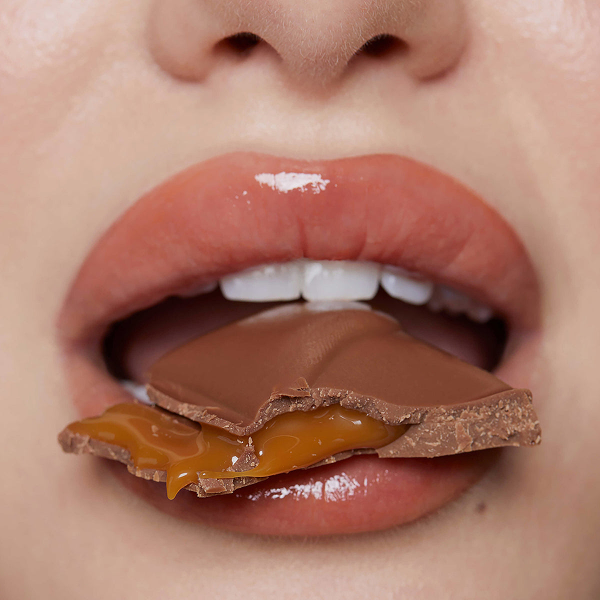 Nahaufnahme von Lippen mit Plumpy Lip Gloss Toffee Brown, die in einen mit Karamell gefülltes Schokostück beißen.