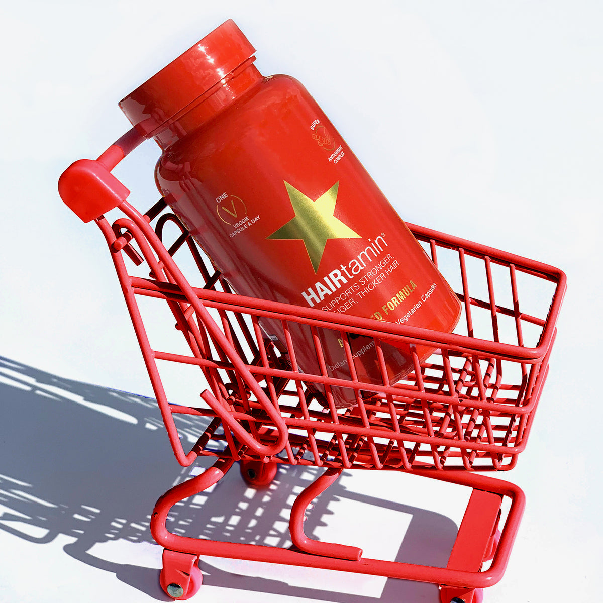 Eine rote Flasche HAIRtamin Advanced Formula 1 Month steht in einem roten Mini-Einkaufswagen vor einem weißen Hintergrund.
