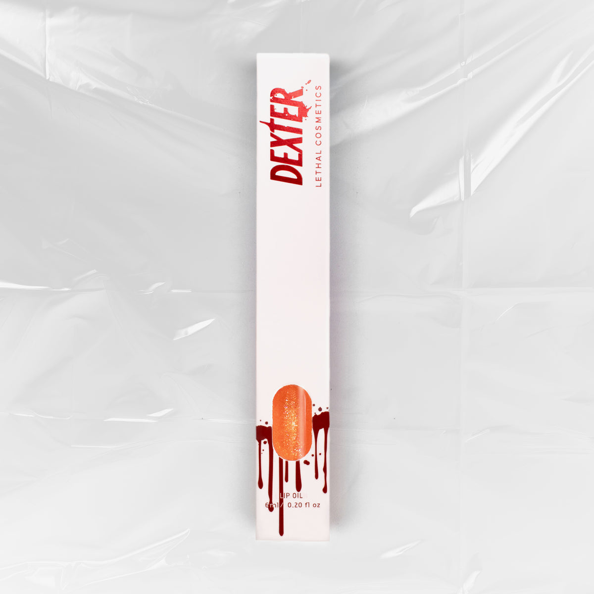 Weiße Schachtel Dexter Lip Oil M99 von Lethal Cosmetics mit Bluttropfen-Design und Logo auf einem Folienhintergrund.