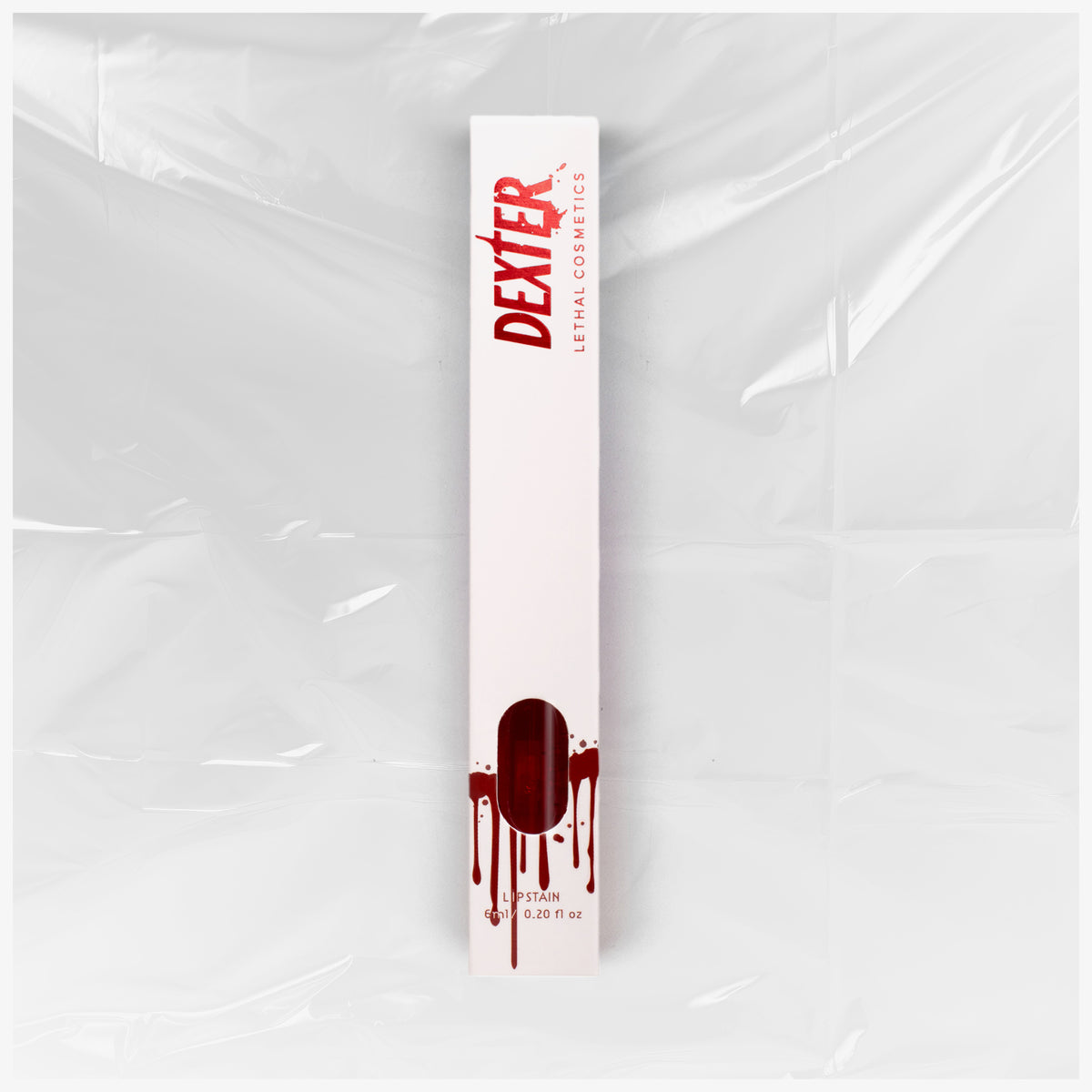Weiße Verpackung des Dexter Lip Stain Born in Blood von Lethal Cosmetics mit rotem Text und Bluttropfen-Design vor einer  Plastikfolie.
