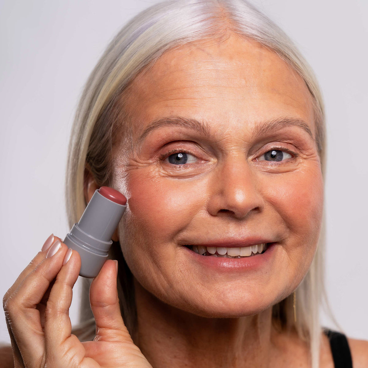 Eine lächelnde ältere Frau trägt Artistick Blush Muse von LH Cosmetics auf ihre Wange auf.