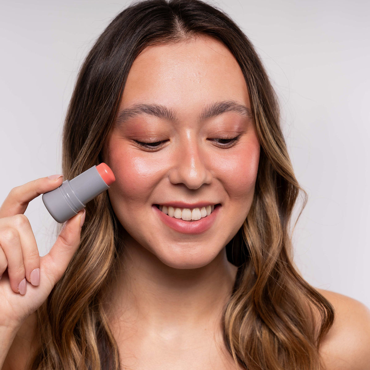 Eine lächelnde Frau trägt vor einem schlichten Hintergrund LH Cosmetics Artistick Blush Sketch in Pfirsich auf ihre Wange auf.