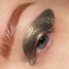 Nahaufnahme eines Auges mit dramatischem Augen-Make-up in funkelndem Olivgold und Schwarz, grafisch über Lid und Augenwinkel hinaus aufgetragen.