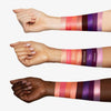 Auf den Unterarmen von drei Armen mit unterschiedlichen Hauttönen sind beschriftete rosa, orange und violette Farbmuster der Bad Side Zodiac Eyeshadow Palette zu sehen.