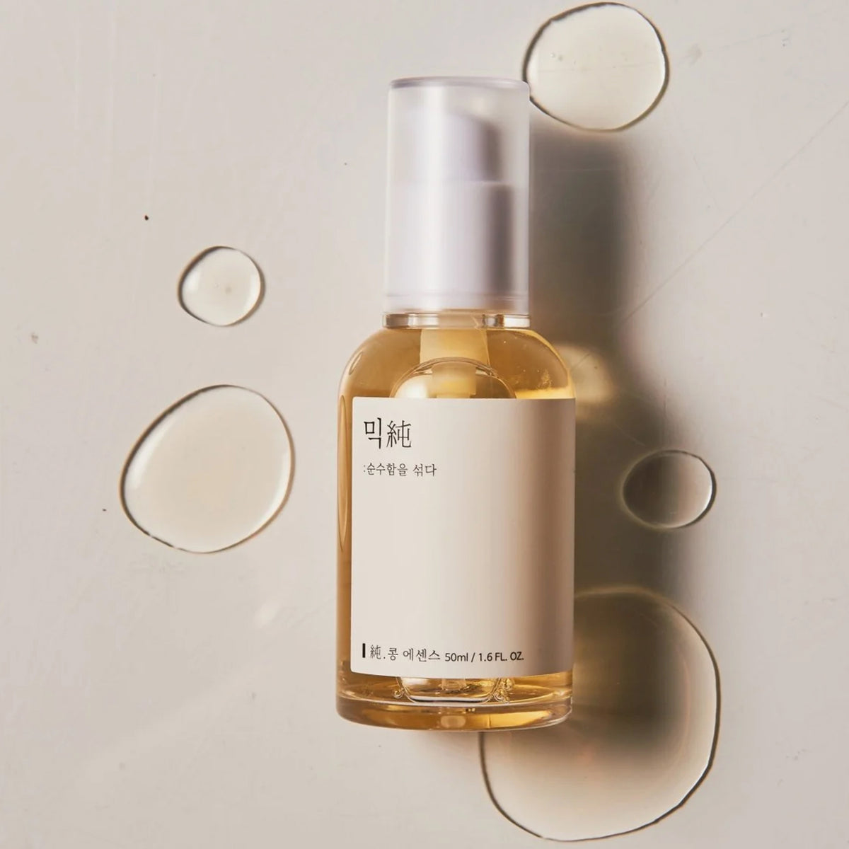 Die Mixsoon Bean Essence in einer klaren Sprühflasche mit weißem Etikett umgeben von Serumtropfen auf einer hellen Oberfläche.