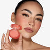 Eine Frau mit kräftigen Augenbrauen hält vor einem weißen Hintergrund das Close-Up Blurring Powder Blush Compact an ihr Gesicht und trägt es auch auf ihren Wangen.