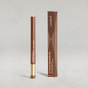 Hellbrauner Lidschatten-Stift mit goldener Kappe von NABLA neben der passenden Umverpackung mit goldenen Designelementen auf hellem Hintergrund.