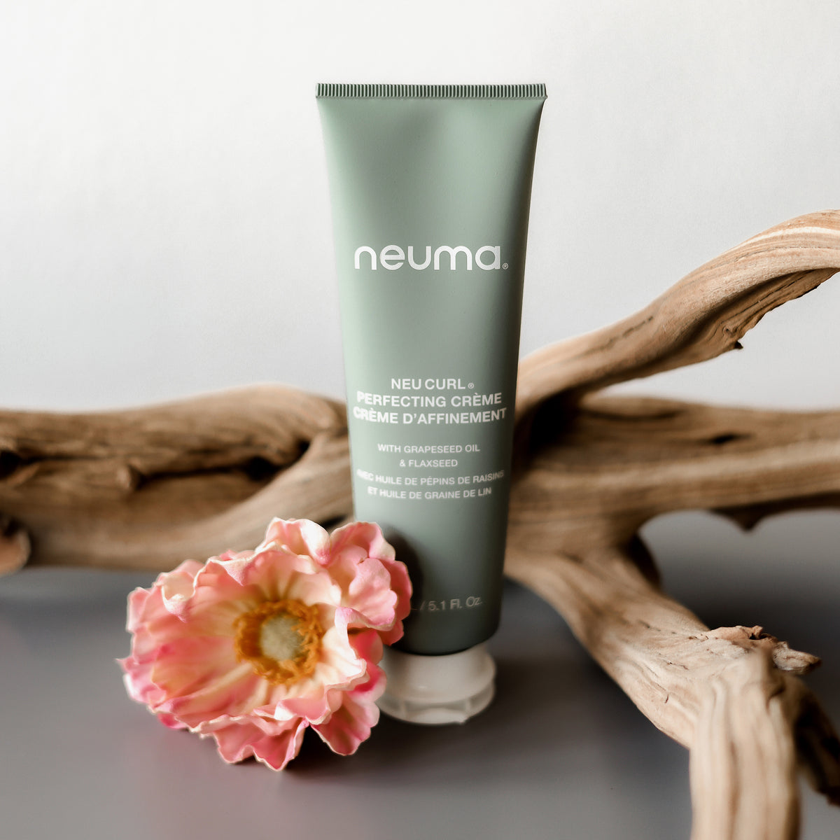 Neuma NEU CURL™ PERFECTING CRÈME in einer grünen Tube wird mit einer rosa Blume und Treibholz auf grauem Untergrund abgebildet.