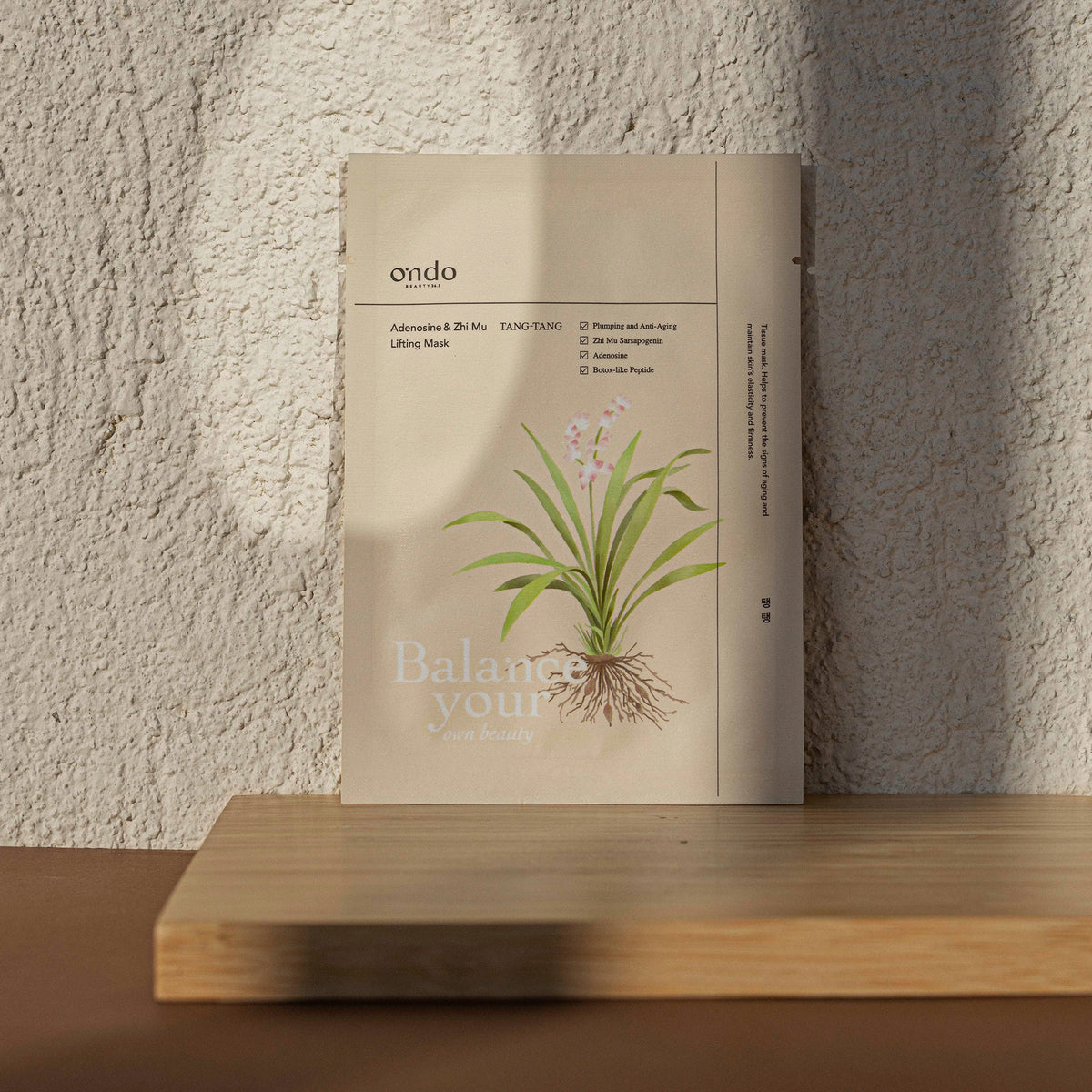 Die Packung „Adenosine & Zhi Mu Lifting Mask Tang-Tang“ mit einer Pflanzenillustration liegt auf einem Holzregal vor einer strukturierten Wand.