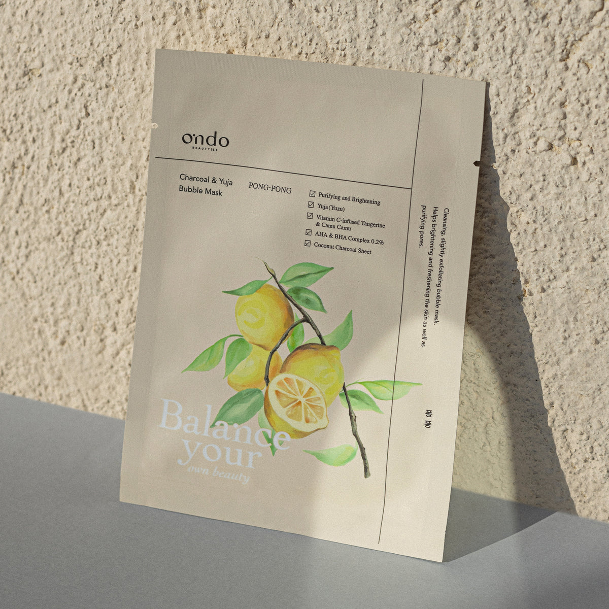 Eine Packung Charcoal & Yuja Bubble Mask Pong-Pong mit Zitronen auf der Vorderseite, lehnt an einer strukturierten Wand.