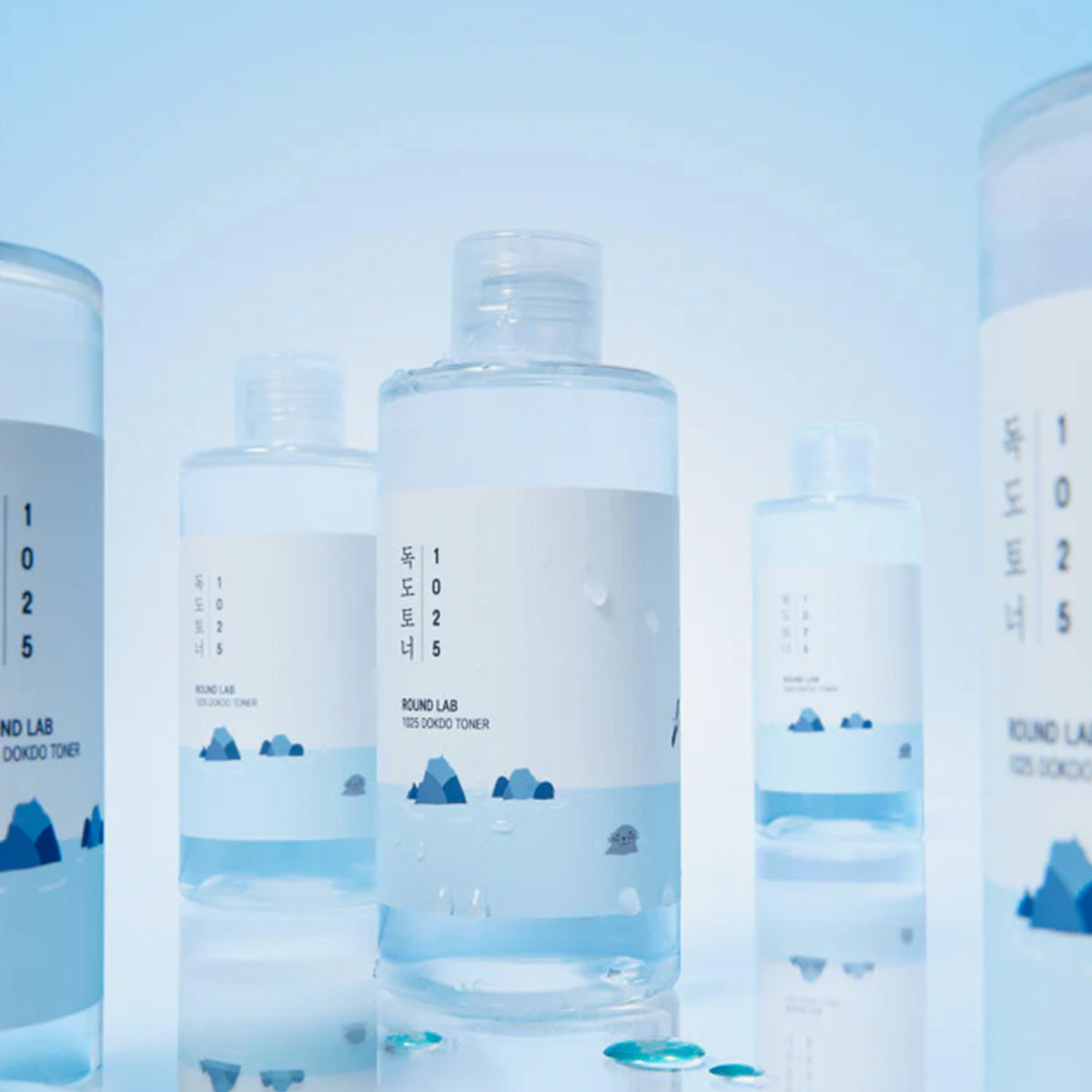 1025 Dokdo-Tonerflaschen mit blauen und weißen Etiketten sind auf einer hellblauen Oberfläche mit Wassertropfen angeordnet.