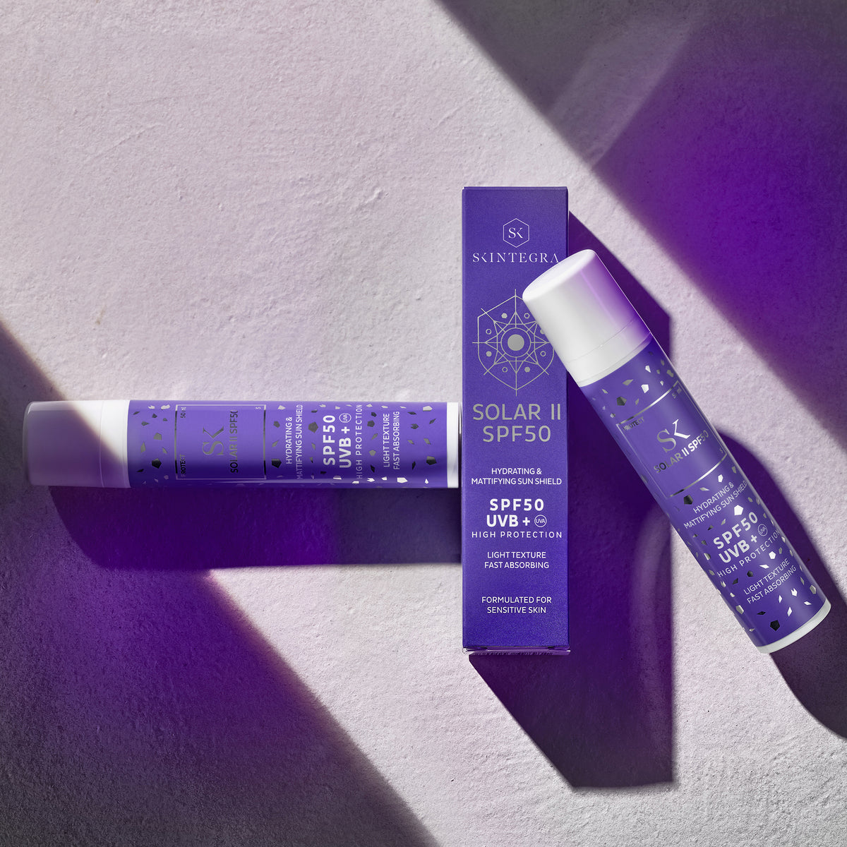 Zwei violette Solar II SPF50-Flaschen und eine Schachtel, auf einer strukturierten Oberfläche unter dramatischer Beleuchtung platziert.