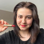 Frau mit schulterlangem dunklem Haar lächelt und hält einen roten Lippenstift in goldener Hülse neben ihr Gesicht.