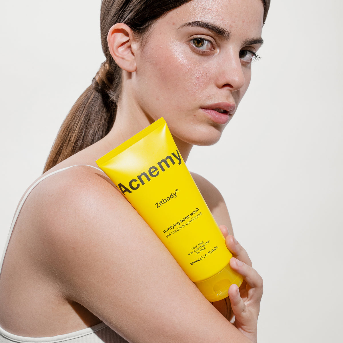 Eine Person hält vor einem neutralen Hintergrund eine gelbe Tube Zitbody zur Bekämpfung von Körperakne in der Nähe ihrer Schulter.
