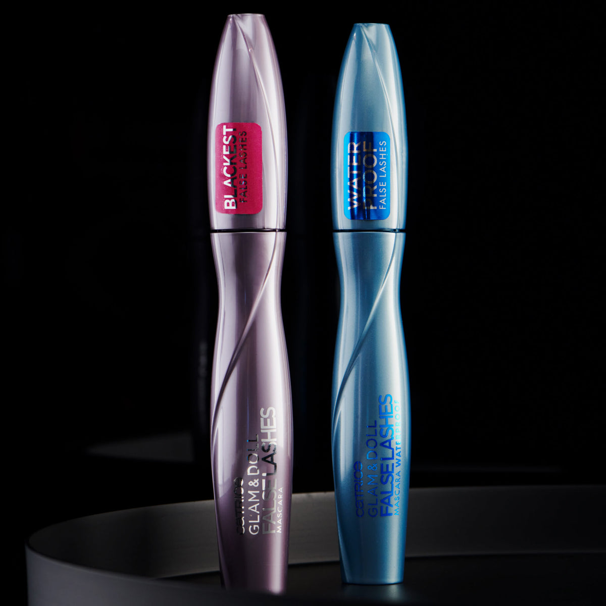 Vor einem dunklen Hintergrund sind zwei Tuben der Glam & Doll False Lashes Mascara abgebildet, eine in Lila, die andere in Blau.