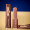 Zwei braune Wonder Woman Butter Bronzer Sticks von Catrice auf beigem Untergrund vor blauem Hintergrund.