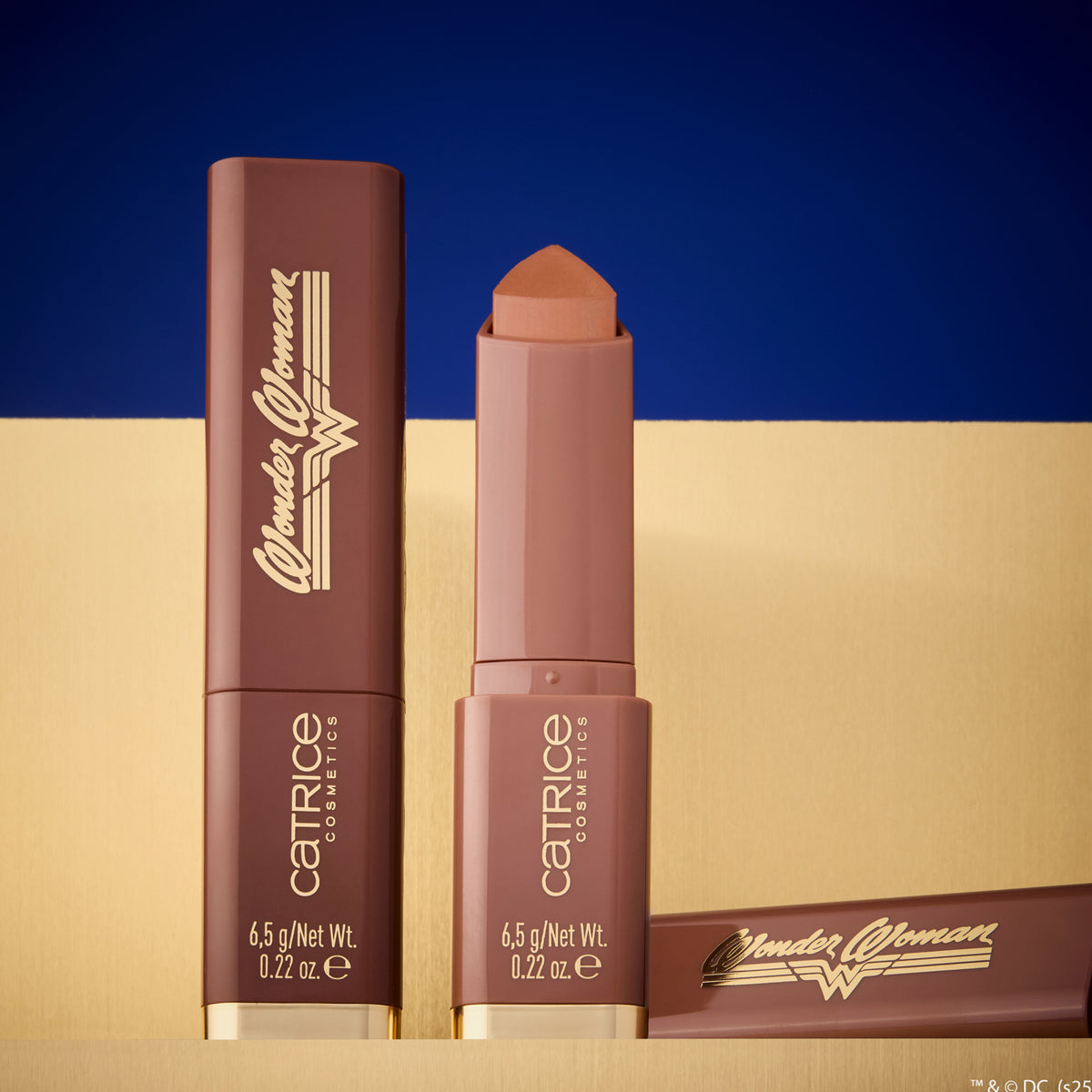 Zwei braune Wonder Woman Butter Bronzer Sticks von Catrice auf beigem Untergrund vor blauem Hintergrund.