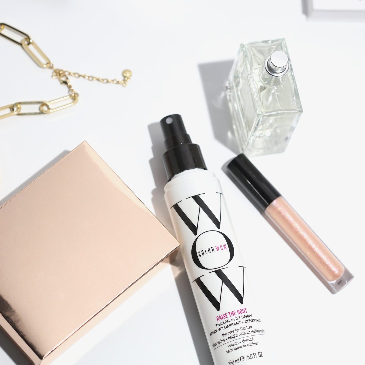 Raise The Root Thicken & Lift Spray neben Lipgloss, Parfümflakon und goldenem Armband auf einer weißen Oberfläche.