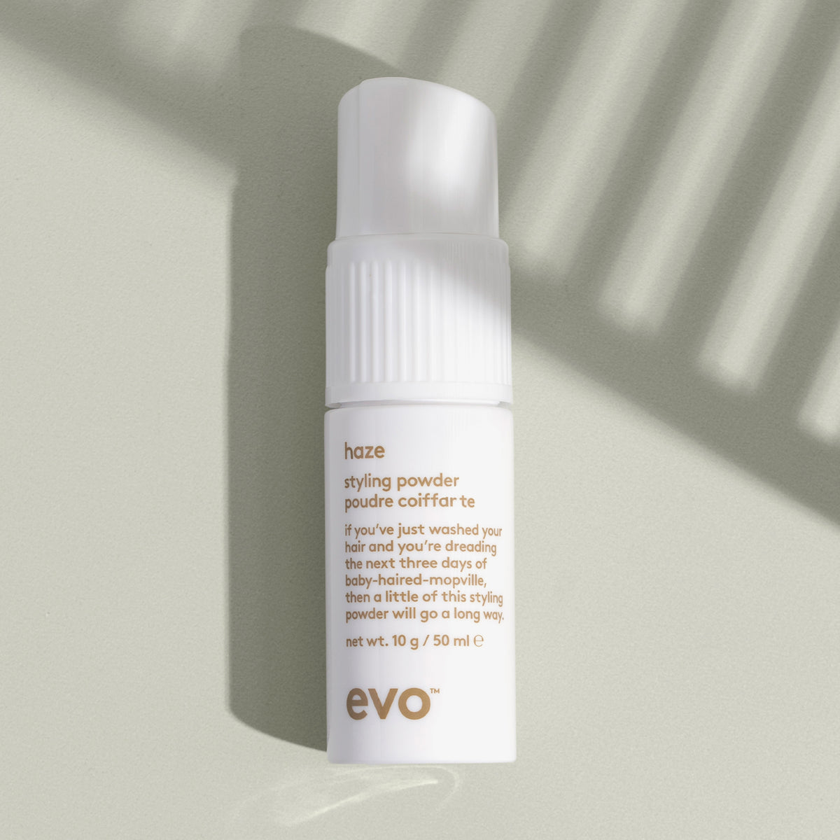Eine weiße Flasche Haze Styling Powder liegt auf einer salbeigrünen Oberfläche, umgeben von weichen Schattenmustern.