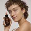 Eine Person hält eine Flasche Vegan Collagen + Organic Oat Barrier & Hydro Toner nah an ihr Gesicht und lächelt leicht.