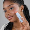 Eine Frau trägt den Blush Stick – 2-in-1 Creme Rouge mit Pinsel Coral auf ihre Wange auf und blickt in die Kamera.