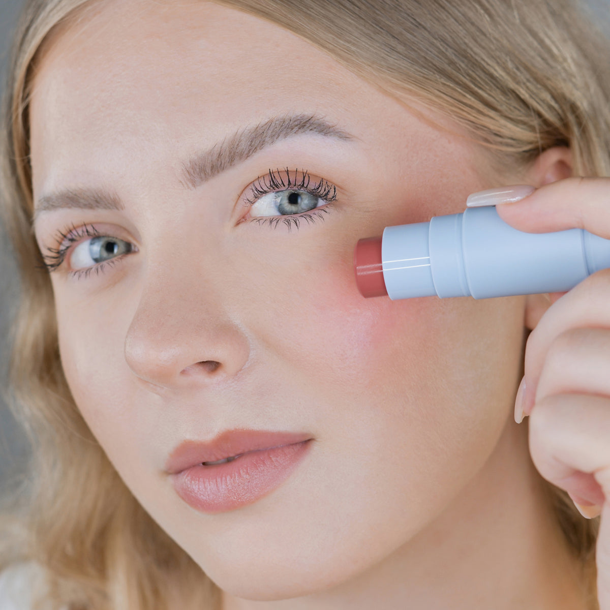 Eine Frau trägt den Blush Stick – 2-in-1 Creme Rouge mit Pinsel Plum auf ihre Wange auf und blickt in die Kamera.