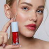 Eine blonde Frau mit glänzendem Make-up hält eine Tube Plumpy Lip Gloss Cherry Red in der Hand.