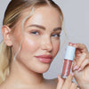 Eine blonde Frau mit glänzendem Make-up hält eine Tube Plumpy Lip Gloss Rose Nude in der Hand.