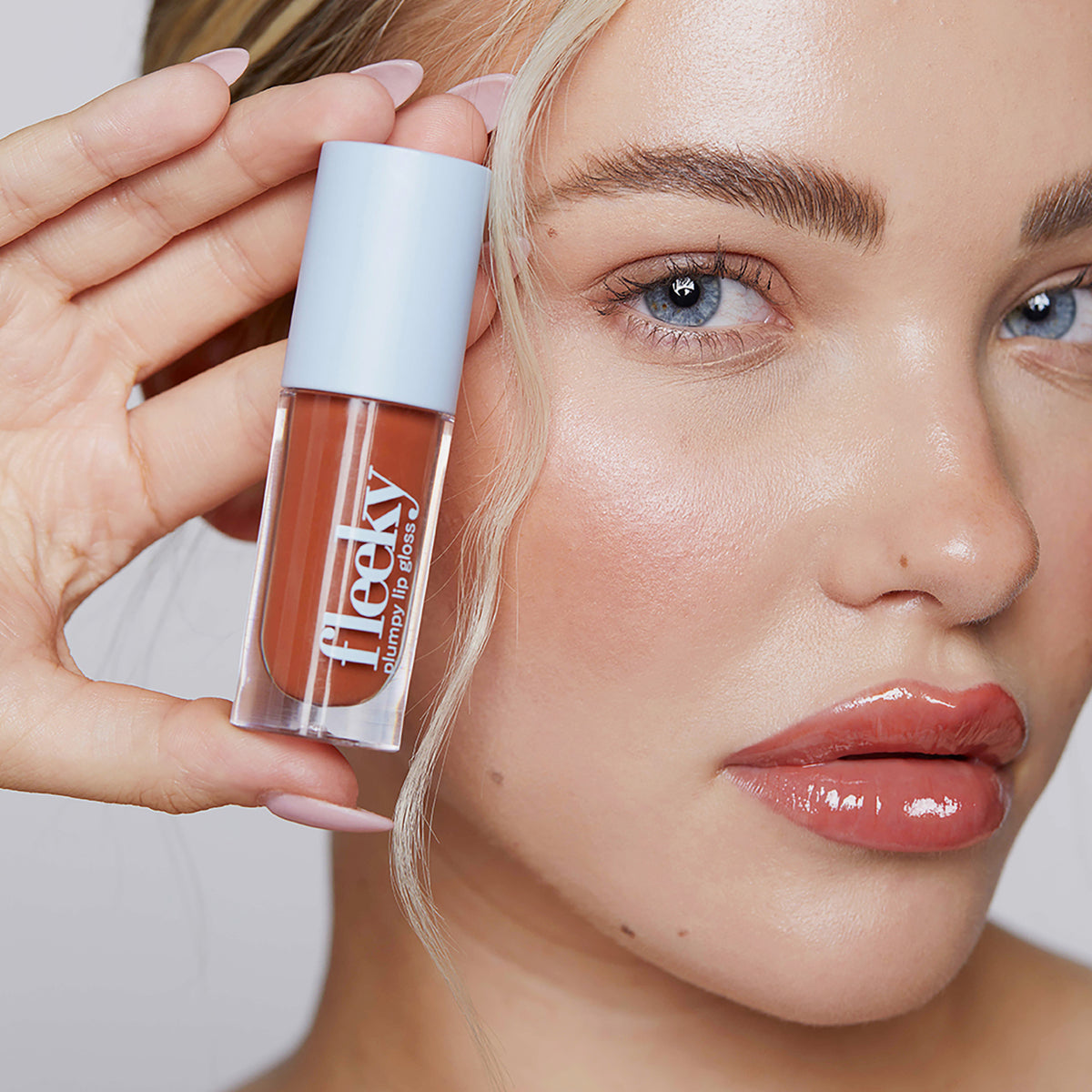Eine blonde Frau mit glänzendem Make-up hält eine Tube Plumpy Lip Gloss Toffee Brown in der Hand.