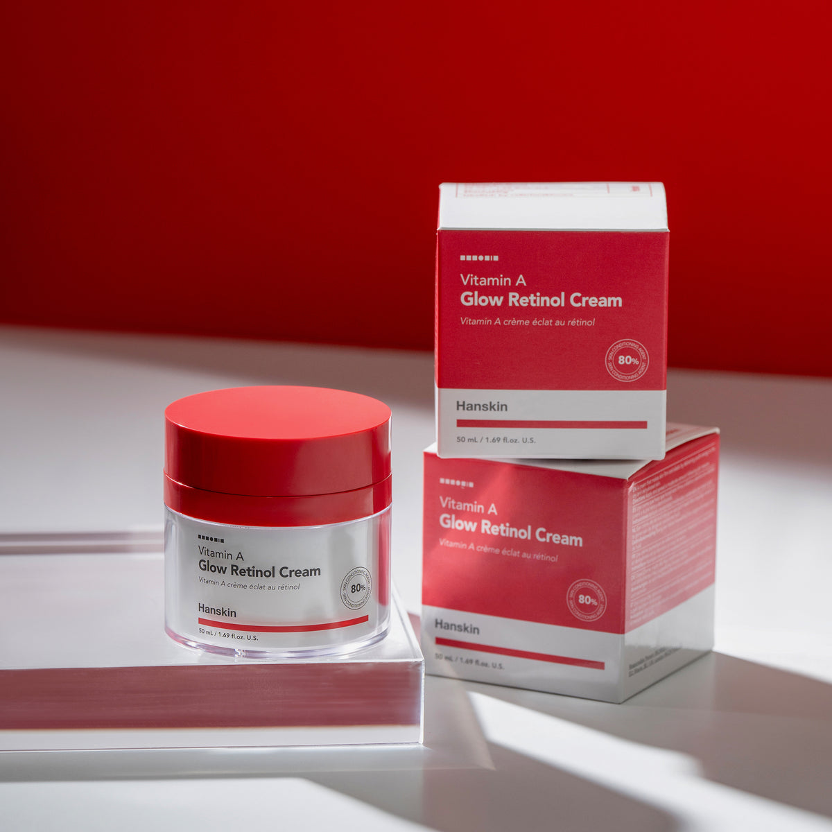 Ein Tiegel und zwei Schachteln Vitamin A Glow Retinol Cream werden auf einer weißen Fläche vor einem roten Hintergrund präsentiert.