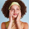 Frau mit lockigem Haar und Stirnband lächelt, während sie ihr Gesicht mit Feel Pure Exfoliation Cleanser wäscht.