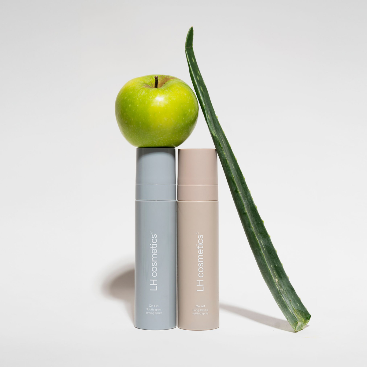 Zwei LH Cosmetics Setting Spray Flaschen, gekrönt von einem grünen Apfel und einem Aloe-Vera-Blatt, vor einem weißen Hintergrund.