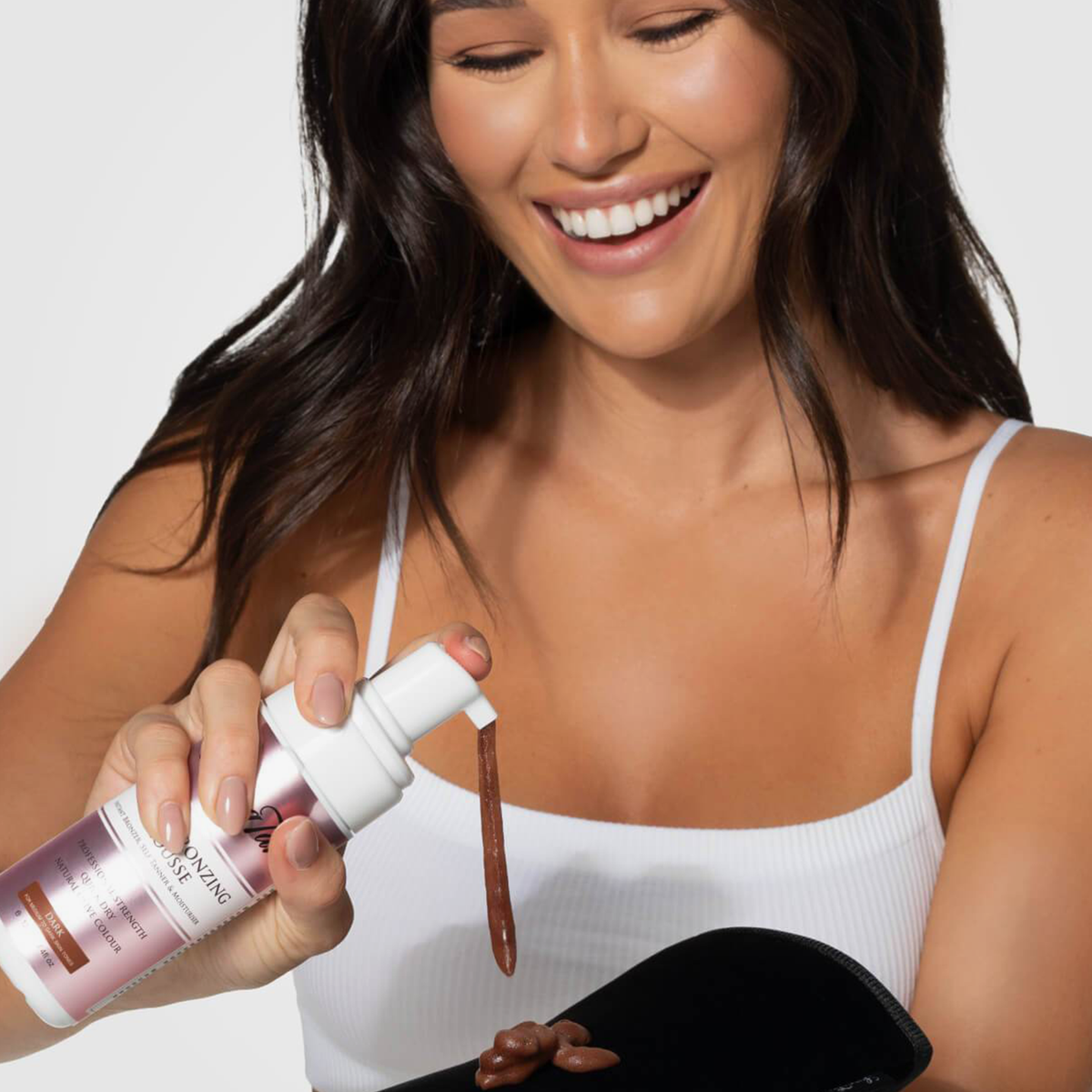 Eine lächelnde Frau in einem weißen Tanktop gibt das Bräunungsmousse Value Bronzing Set von Loving Tan aus der Flasche auf einen Handschuh.